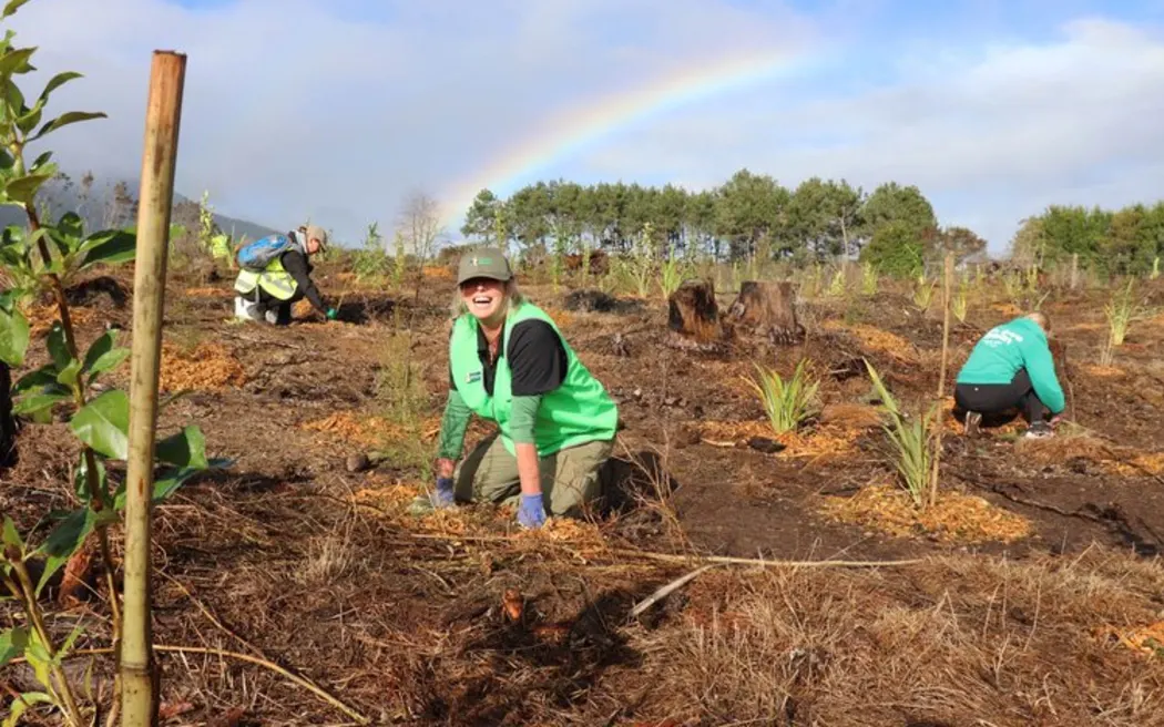 A woman smiling broadly while kneeling next to seedlings in a planting site. She is wearing bright green with a green cap and gardening gloves. There are two people kneeling in the background behind her, planting trees, and a rainbow stretches across the cloudy sky.