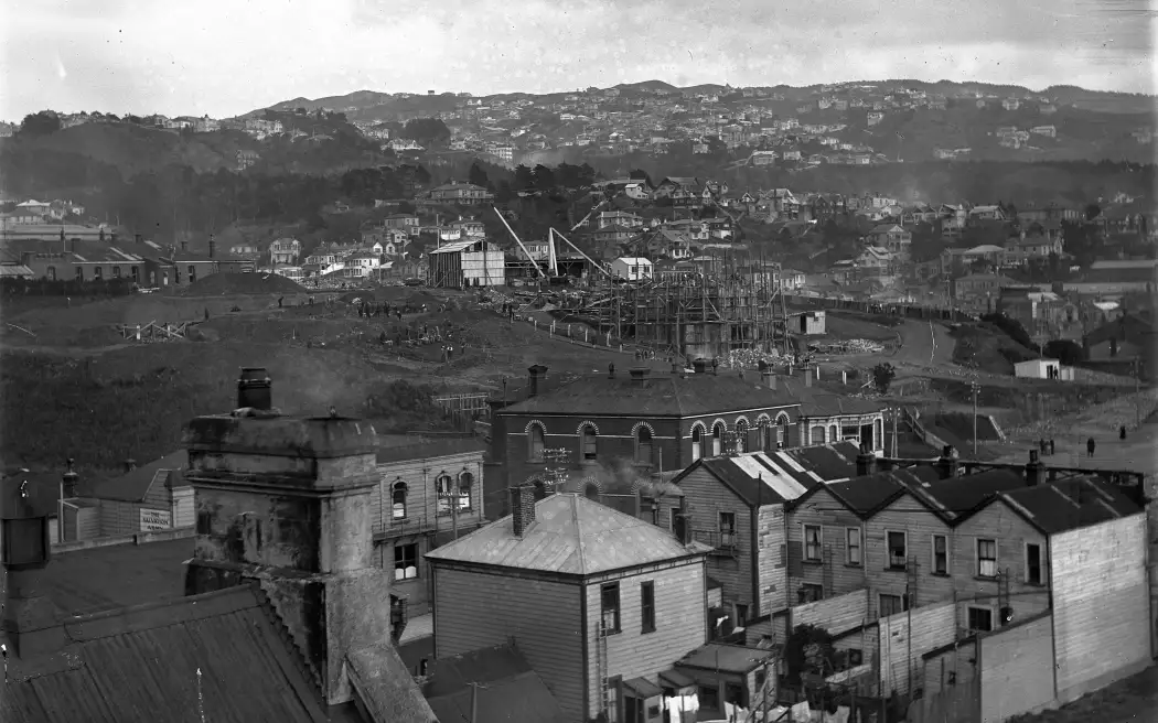 Looking west over Mt Cook in 1931. Shows the police barracks in the middle ground (brick building, arched windows), the back of houses on Tory Street (right) and the carillon under construction. Mt Cook, Wellington, Carillon under construction.
