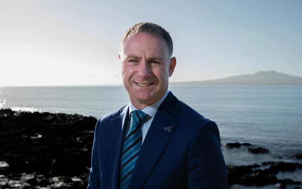 New Zealand: Khí thải hàng không, hàng hải có được đưa vào mục tiêu Net Zero 2050? 2 Climate Change minister Simon Watts, wearing a blue suit, stands in bright sunlight near a rocky shore, with Rangitoto Island in the background.