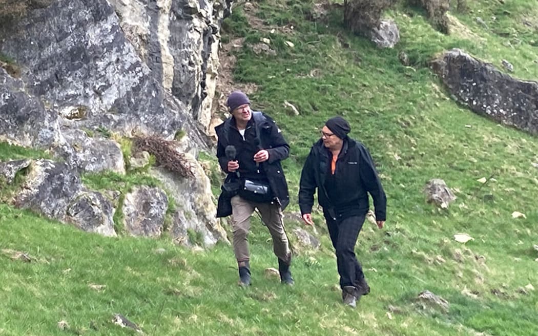 Country Life producer Cosmo Kentish-Barnes carrying recording gear walking up a grassy slope with a farmer against a backdrop of limestone rocks around the Ōpihi River in Canterbury