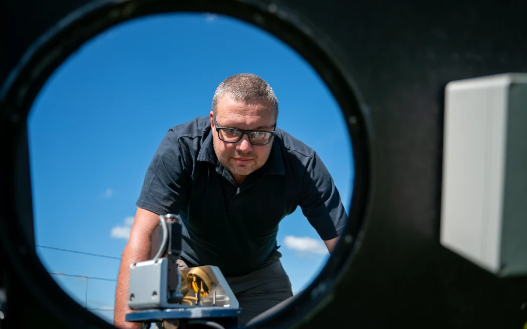 A person wearing glasses and a dark shirt is seen through a circular opening, leaning forward and looking intently at a piece of equipment. The background features a clear blue sky with some clouds.