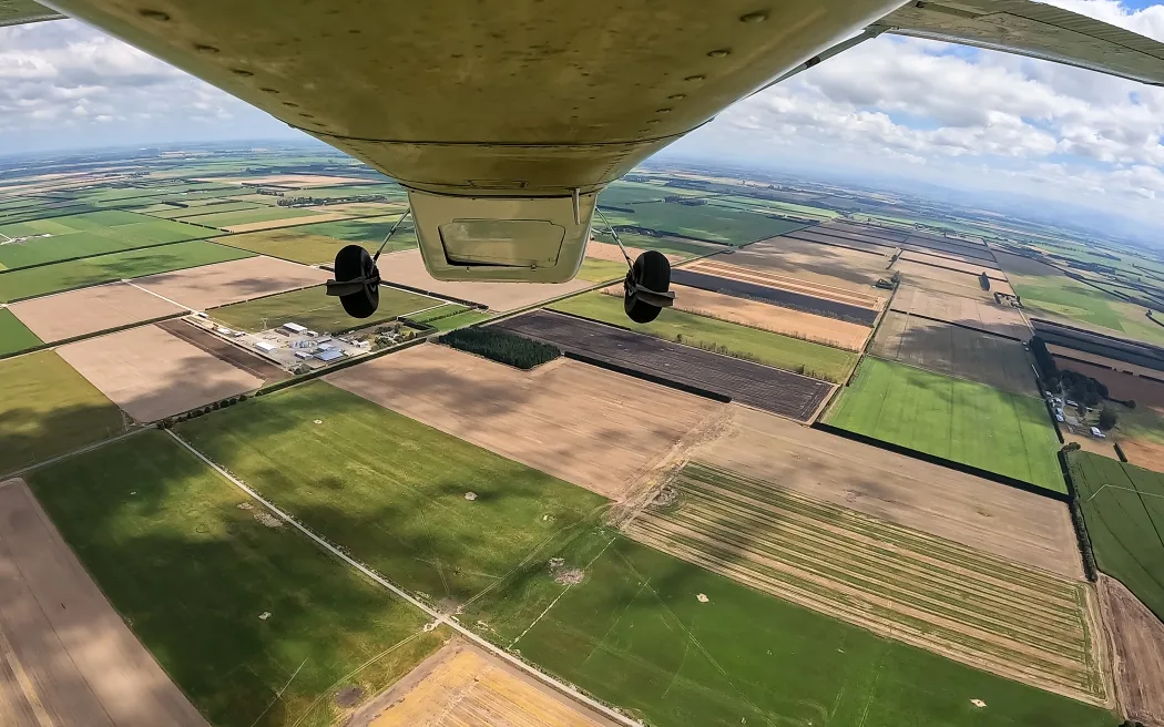 Aerial view from underneath an airplane showing a patchwork of agricultural fields in different shades of green and brown. The horizon is visible in the distance under a partly cloudy sky.