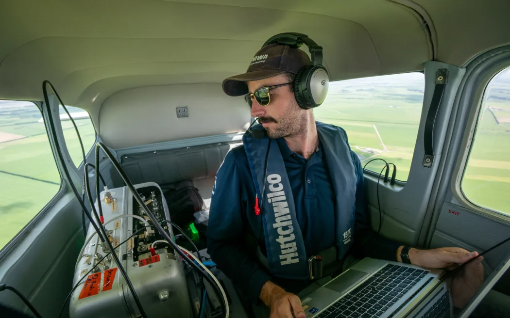 A person seated inside a small aircraft, wearing headphones and sunglasses, holds a laptop and looks out the window. The aircraft interior features various equipment, cables, and a control panel with switches. Outside the window, green fields and farmland are visible below.