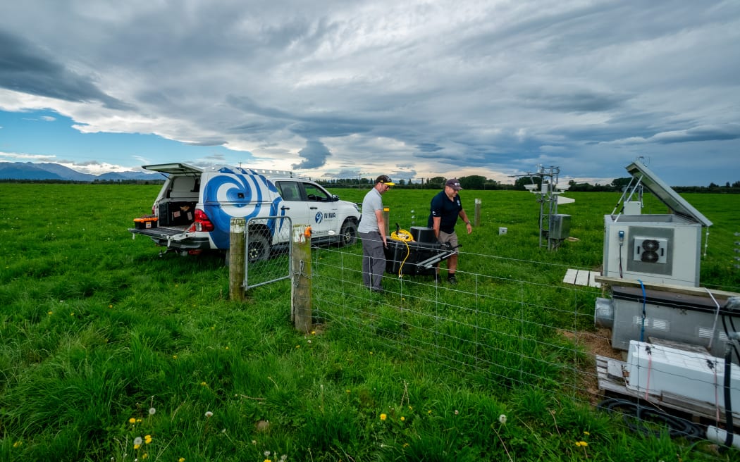 Two individuals carrying scientific equipment in a grassy field. A white vehicle with the 