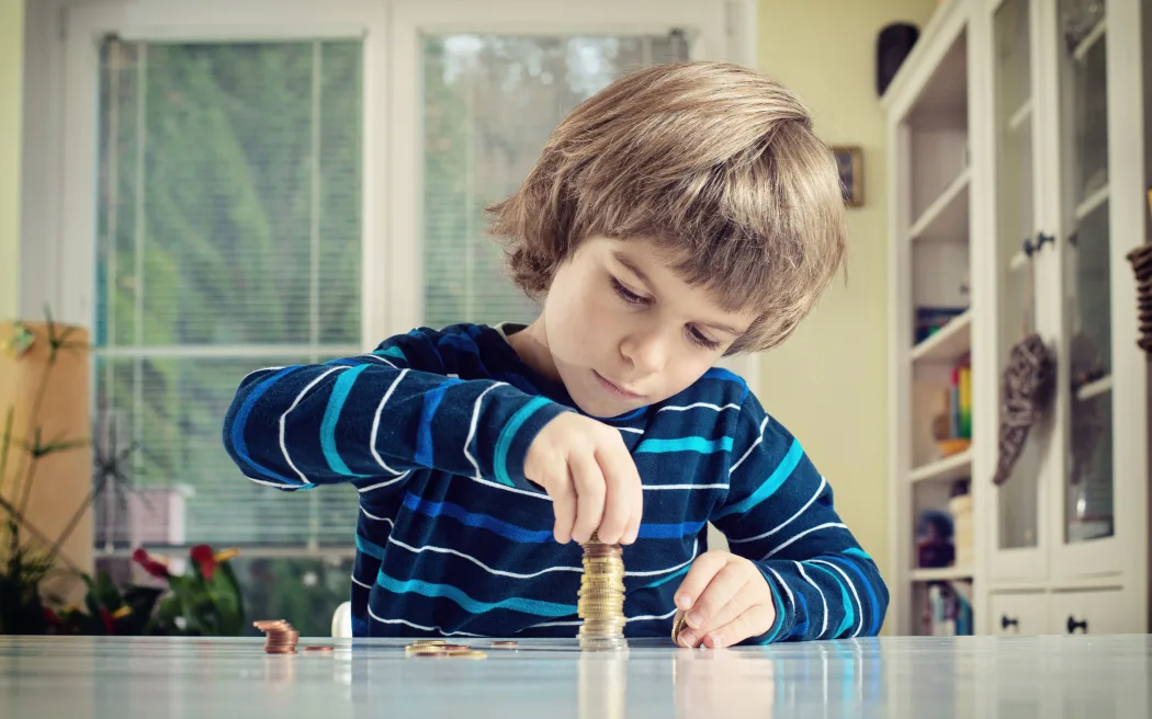 Bí quyết giúp con bạn thành thạo tài chính từ nhỏ 44 A photo of a little boy making stack of coins, counting money at table. Learning financial responsibility and planning savings concept.