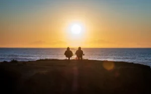 A couple sit together on fold out chairs watching a sunset over the sea.