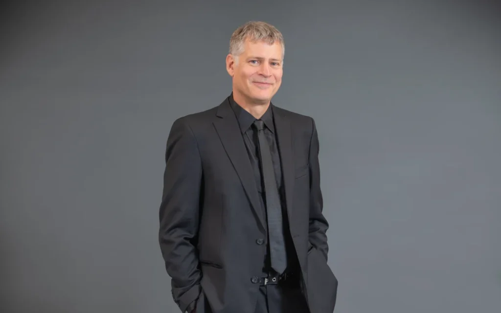 Cải tổ giáo dục New Zealand: Bão tố phản đối hé lộ rạn nứt sâu sắc 1 Michael stands in front of a grey backdrop wearing a grey suit, with his hands tucked into his pockets. He smiles.