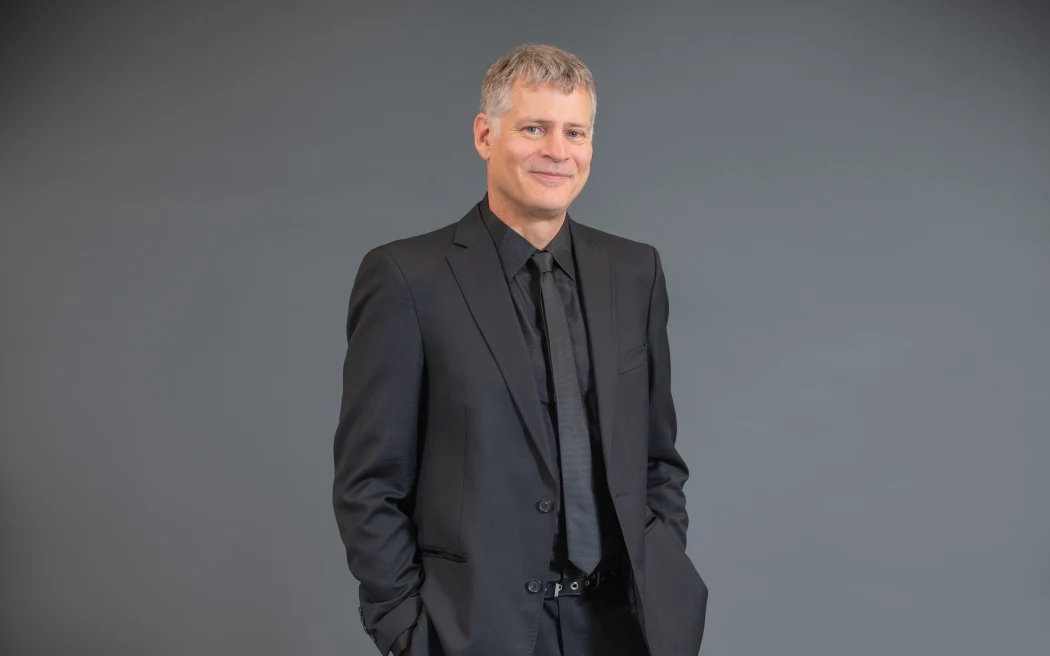 Michael stands in front of a grey backdrop wearing a grey suit, with his hands tucked into his pockets. He smiles.