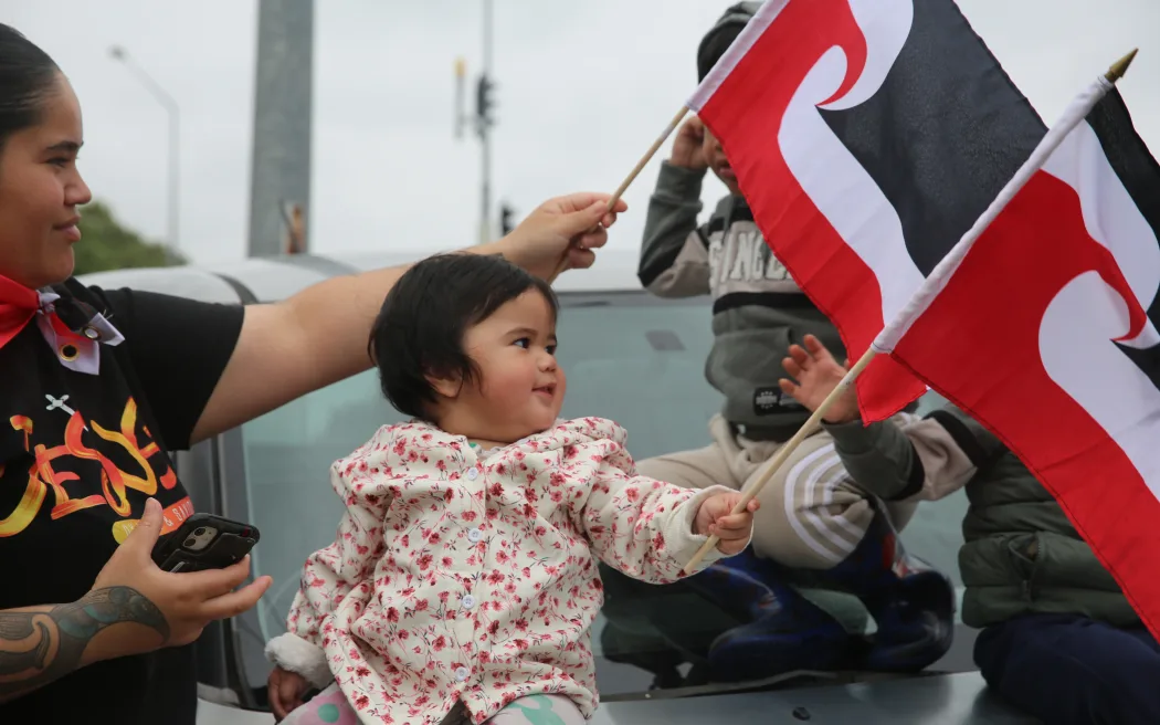Pēpi waves her tinorangatiratanga flag with her mum and siblings at the hīkoi in Tāmaki Makaurau.