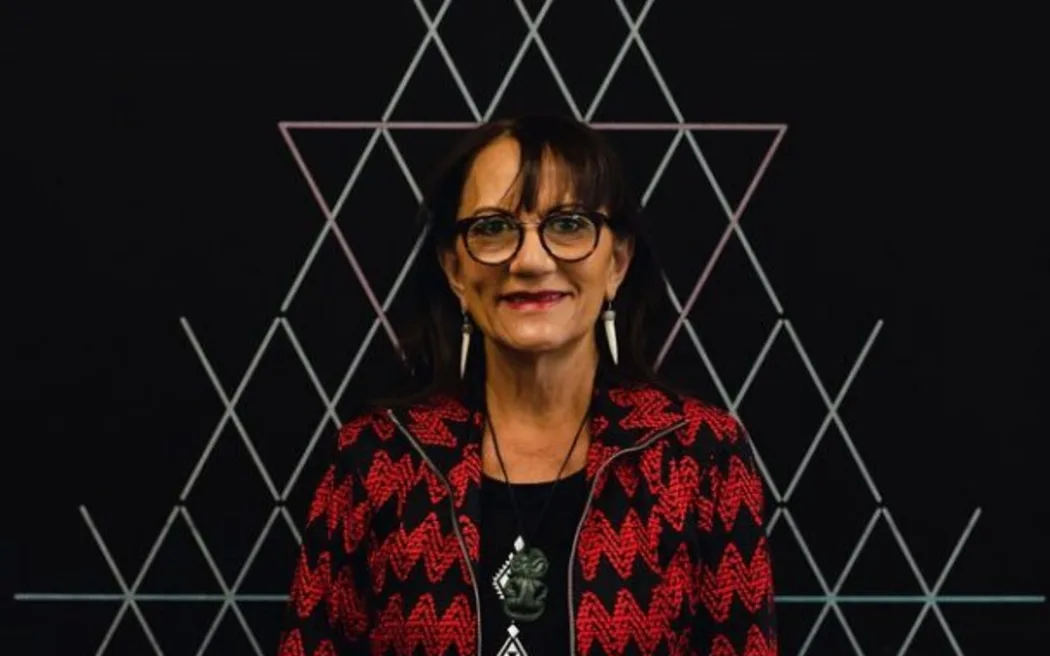 Te Puea smiles at the camera. She stands in front of a black wall designed with white tukutuku panelling.